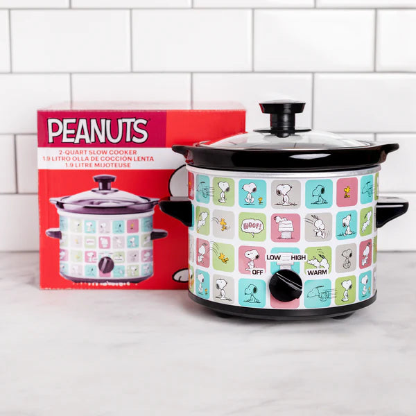 Slow cooker with Peanuts character design next to its packaging on a white tiled wall background