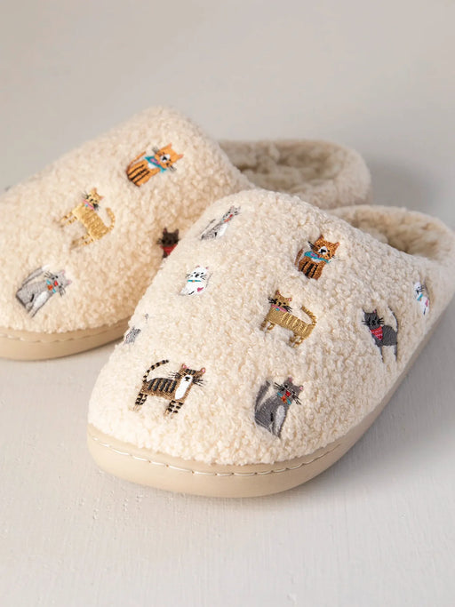 Pair of cream-colored slippers with embroidered cat designs on a light gray background