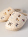 Pair of cream-colored slippers with embroidered cat designs on a light gray background