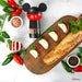 Wooden board with baguette, tomatoes, mozzarella, and a Mickey Mouse-themed pepper grinder on a white background.