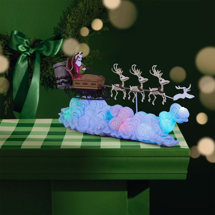 Decorative scene with Santa Claus in a sleigh pulled by skeleton reindeer on a cloud, set against a green checkered tablecloth.