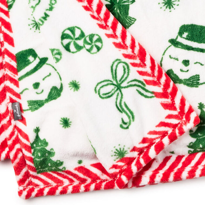 A close-up of a white plush blanket with a festive Christmas print featuring green snowmen, candy canes, pine trees, and a red-and-white striped border.
