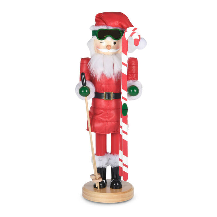 A festive wooden nutcracker styled as Santa Claus skiing, wearing a red suit, green goggles, and holding a red-and-white striped ski pole, standing on a round wooden base against a white background.