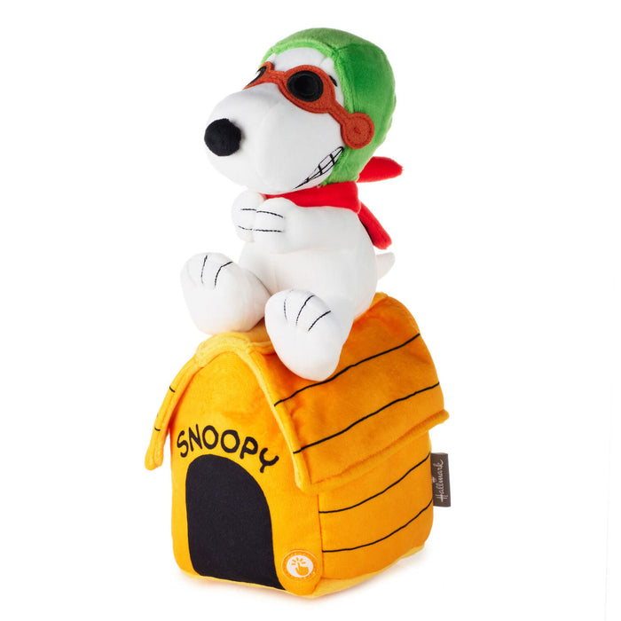 A plush Snoopy figure wearing a green hat and red scarf, sitting on top of a yellow dog house with black stripes and the word 'SNOOPY' on the front, photographed on a white background.