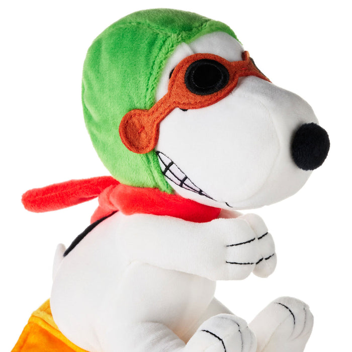 A plush toy of Snoopy dressed as a superhero, wearing a green helmet, orange mask, red scarf, and yellow shorts, set against a plain white background.