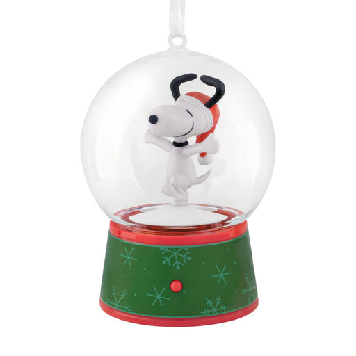 A Snoopy snow globe ornament featuring Snoopy hugging his dog friend inside a clear glass globe, mounted on a green and red holiday-themed base with snowflake patterns.