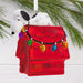 A Snoopy doghouse Christmas ornament with a white dog figure on top, decorated with a red house body, hanging white ribbon, and colorful string lights, shown against green pine branches and a white background.