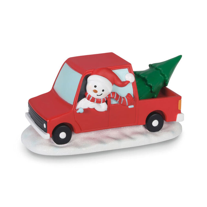 A small holiday figurine of a red pickup truck carrying a green Christmas tree, with a smiling snowman wearing a red Santa hat and striped scarf sitting in the driver's seat, all mounted on a white base against a plain white background.