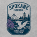 A square graphic print featuring a vintage-style shield emblem with the words 'SPOKANE STRONG' at the top and 'REBUILDING TOGETHER' at the bottom. The illustration includes a bridge, tower, trees, waterfall, and purple flowers on a light gray patterned background.