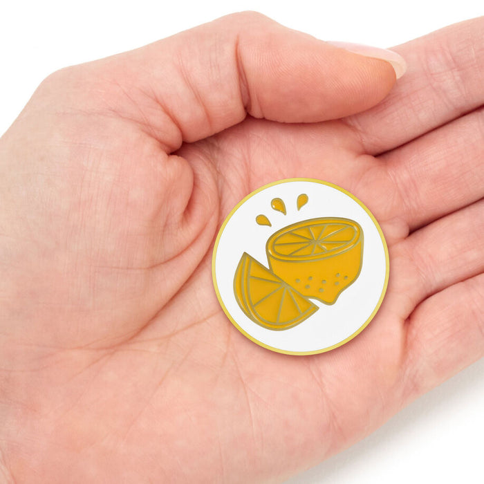 A small round enamel pin featuring a yellow lemon slice illustration on a white background, held in the palm of a hand.