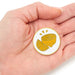 A small round enamel pin featuring a yellow lemon slice illustration on a white background, held in the palm of a hand.