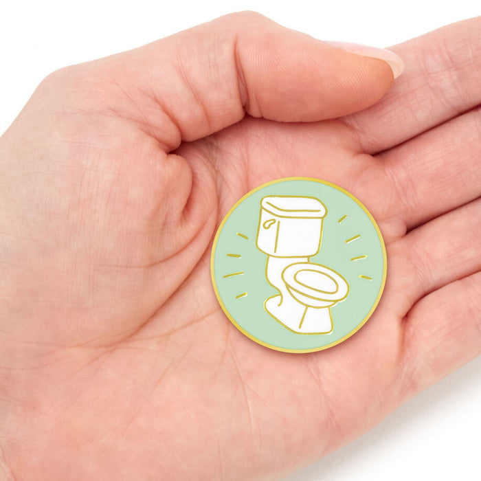 A small round enamel pin featuring a white toilet illustration on a light green background with yellow accents, held in the palm of a hand.