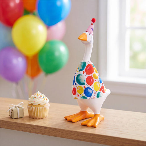 A white goose figurine wearing a colorful party hat and a polka-dot balloon dress stands on a wooden table next to a small wrapped gift box and a cupcake. Blurred balloons and a bright window appear in the background.
