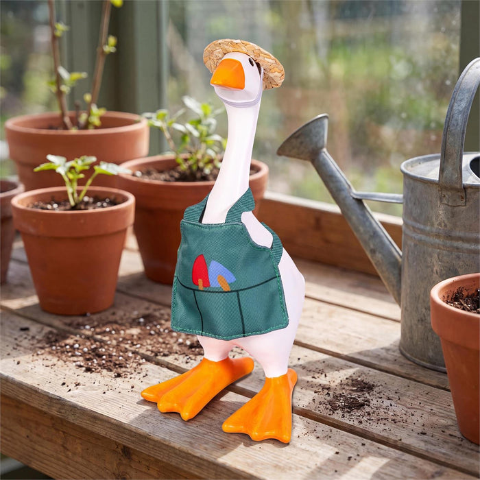 A decorative garden goose figurine wearing a teal gardening apron with colorful pockets and a straw hat, standing on a wooden table beside terracotta pots and a metal watering can in a garden setting.