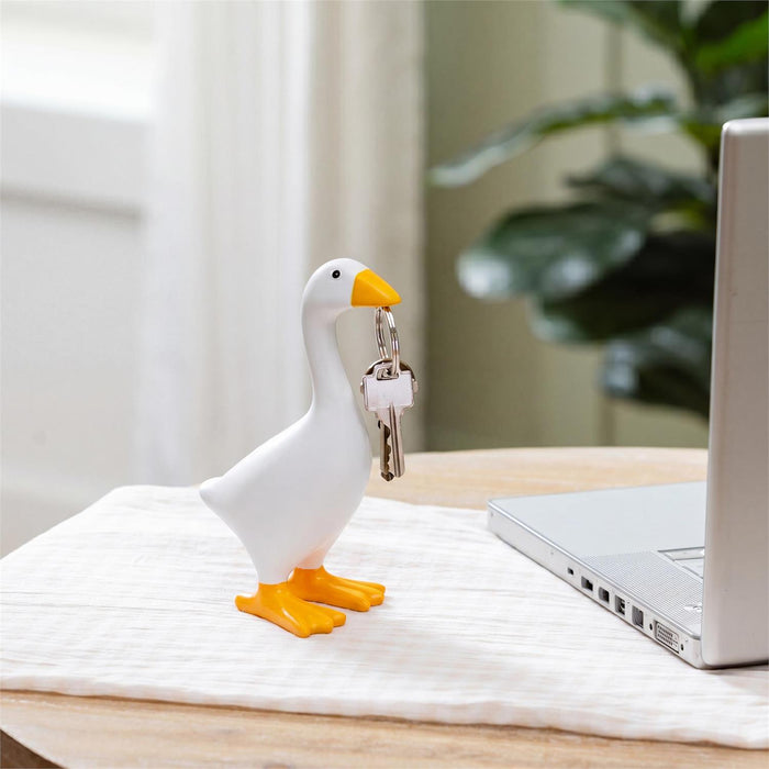 A white goose-shaped key holder with yellow beak and feet, holding a set of keys, placed on a white textured cloth on a wooden table.