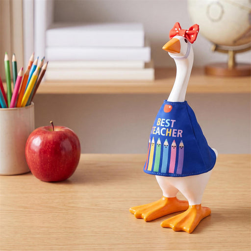 A decorative goose figurine wearing a blue shirt that reads 'BEST TEACHER' with colorful pencil graphics, standing on a wooden desk next to a red apple and pencils.