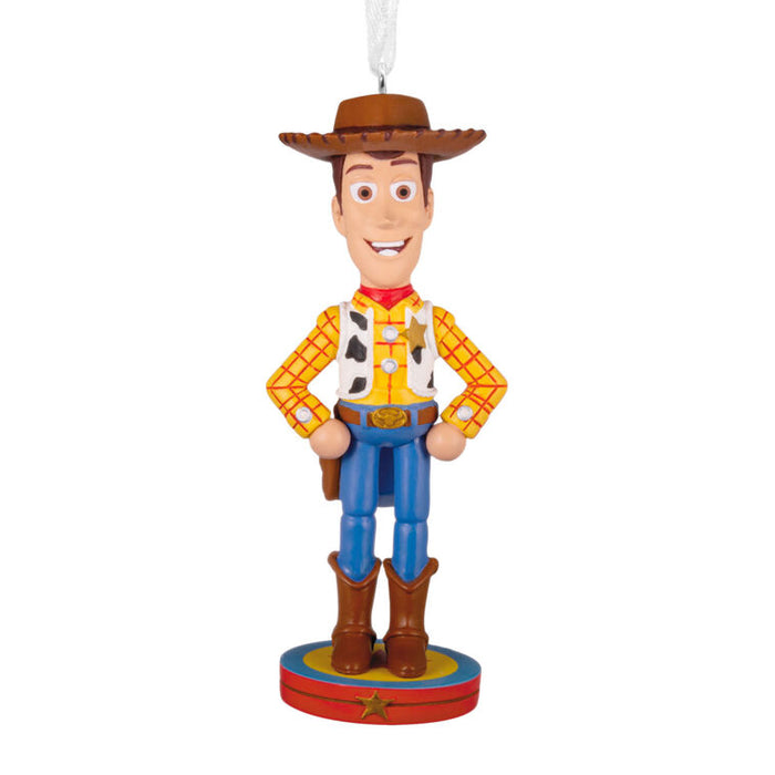 A Woody the Cowboy character ornament from Toy Story, featuring his signature yellow plaid shirt, brown hat and boots, blue pants, and red vest details, standing on a circular base with a hanging loop.