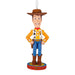 A Woody the Cowboy character ornament from Toy Story, featuring his signature yellow plaid shirt, brown hat and boots, blue pants, and red vest details, standing on a circular base with a hanging loop.