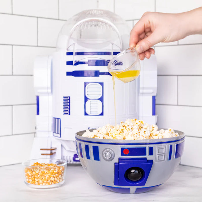 R2-D2 popcorn maker with a hand pouring oil into it, set against a white tiled wall.