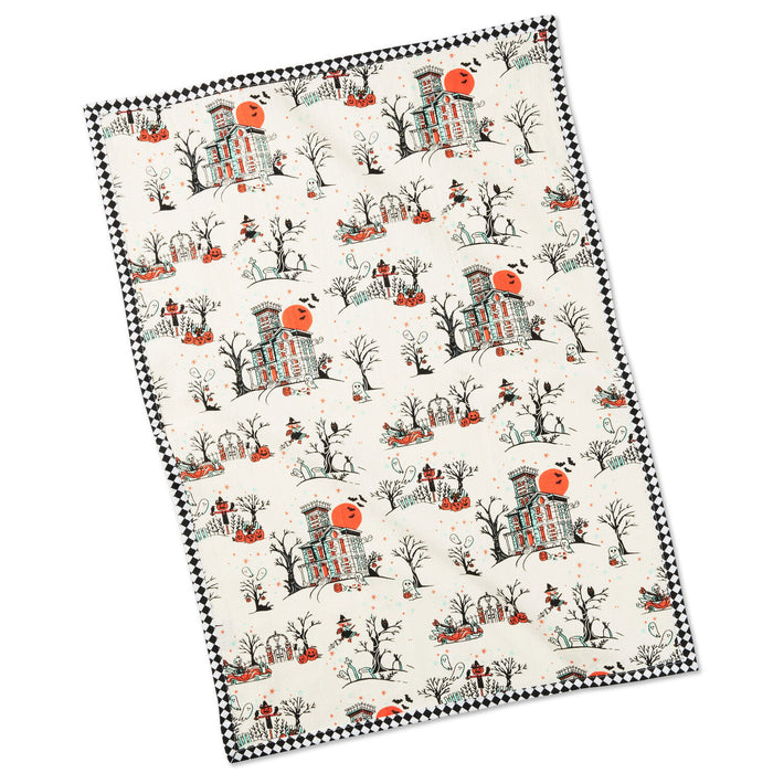 A rectangular fabric item, likely a tea towel or blanket, featuring a repeating Halloween pattern of haunted houses, pumpkins, bats, and trees on an off-white background, finished with a black and white checkered border.