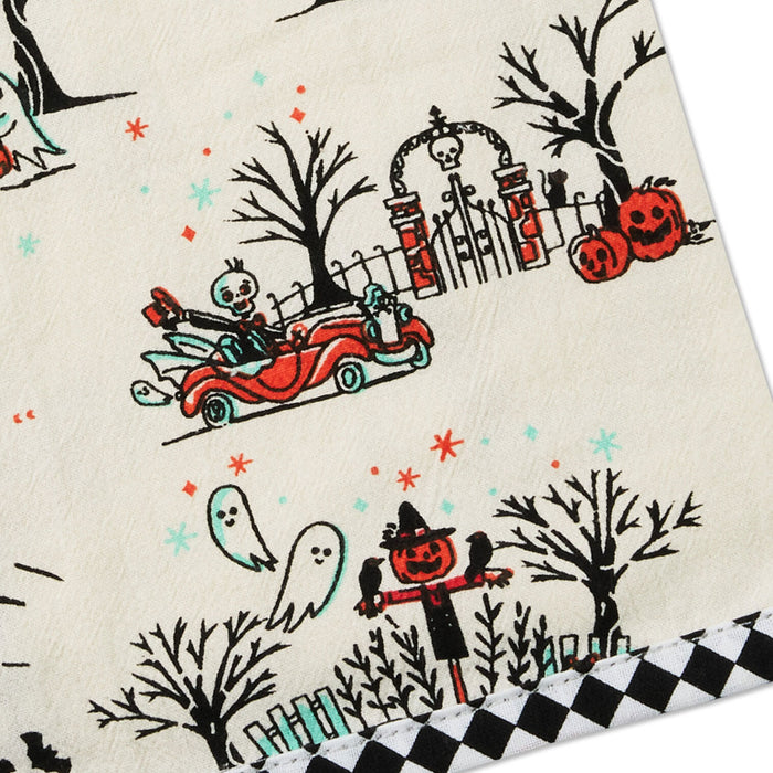 A close-up of a Halloween-themed fabric or paper product featuring a cream background with printed black trees, a red convertible, skeleton driver, pumpkins, ghosts, and a black-and-white checkered border.