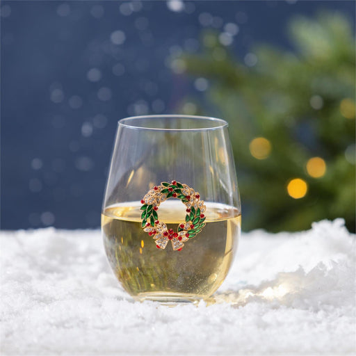A clear stemless wine glass filled with white wine sits on white snow, with a small holiday wreath brooch resting on the rim. The background is a soft, blurred Christmas tree and dark blue sky with falling snow.