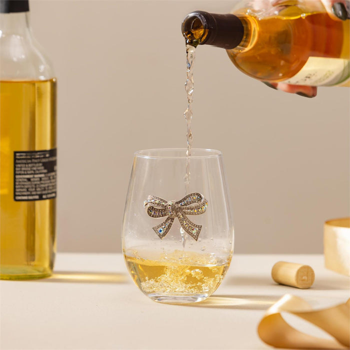 A stemless wine glass with a sparkling rhinestone bow on the front, shown with white wine being poured into it from a bottle. A second bottle stands in the background, along with a cork and a strip of paper on a light beige surface.