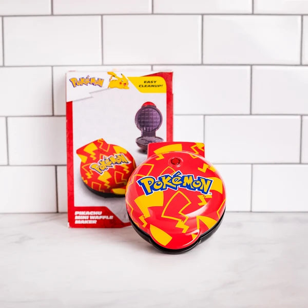 A red and yellow Pokémon Peachu Mini Squebble Ball toy in the foreground with its original packaging box standing behind it on a white marble surface against a white subway tile wall.