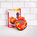 A red and yellow Pokémon Peachu Mini Squebble Ball toy in the foreground with its original packaging box standing behind it on a white marble surface against a white subway tile wall.