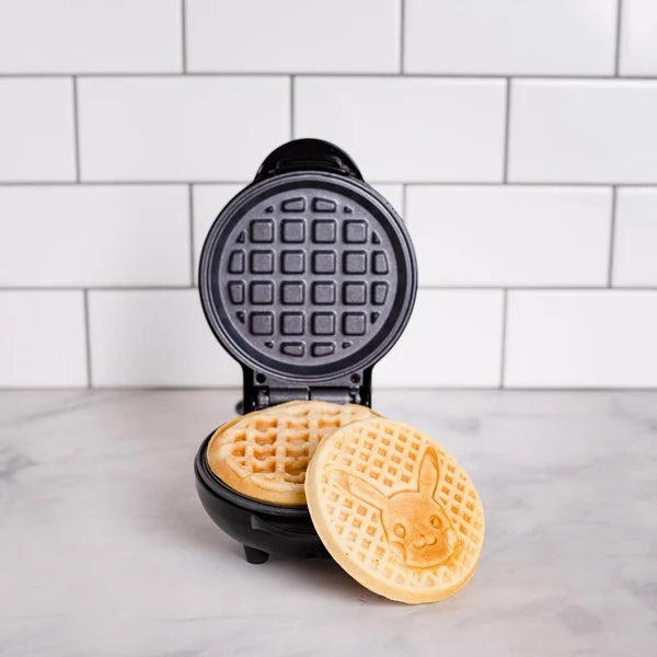 A black mini waffle maker shown open on a marble countertop with white subway tile in the background. Two golden-brown waffles are visible inside and one waffle is leaning against the outside of the lower plate, featuring a bunny face design.