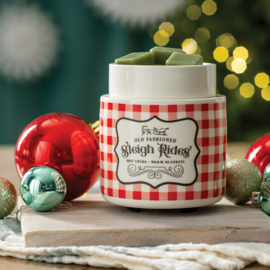 A ceramic hot cocoa and warm blanket maker jar labeled 'Old Fashioned Sleigh Rides' with a red-and-white gingham pattern, shown on a wooden board with holiday ornaments and a blurred Christmas tree background.