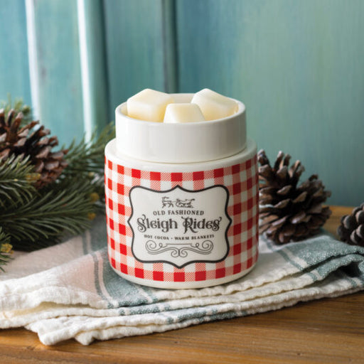 A ceramic jar candle labeled 'Old Fashioned Sleigh Rides' with a red and white gingham pattern and white wax melts visible at the top, styled on a wooden surface with a cloth, pinecones, and evergreen branches against a teal background.