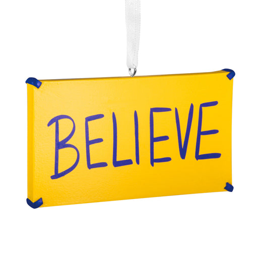 A rectangular yellow hanging ornament or sign with the word “BELIEVE” written across it in large blue letters, suspended by a white ribbon against a plain white background.