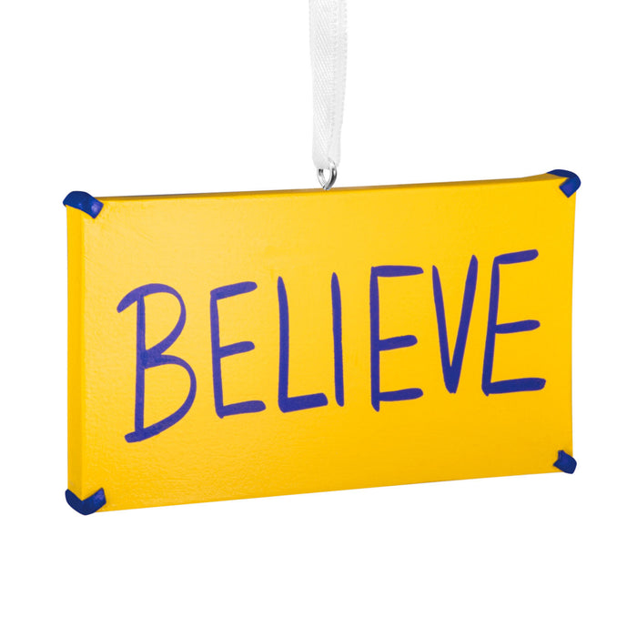 A rectangular yellow hanging ornament or sign with the word “BELIEVE” written across it in large blue letters, suspended by a white ribbon against a plain white background.