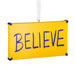 A rectangular yellow hanging ornament or sign with the word “BELIEVE” written across it in large blue letters, suspended by a white ribbon against a plain white background.