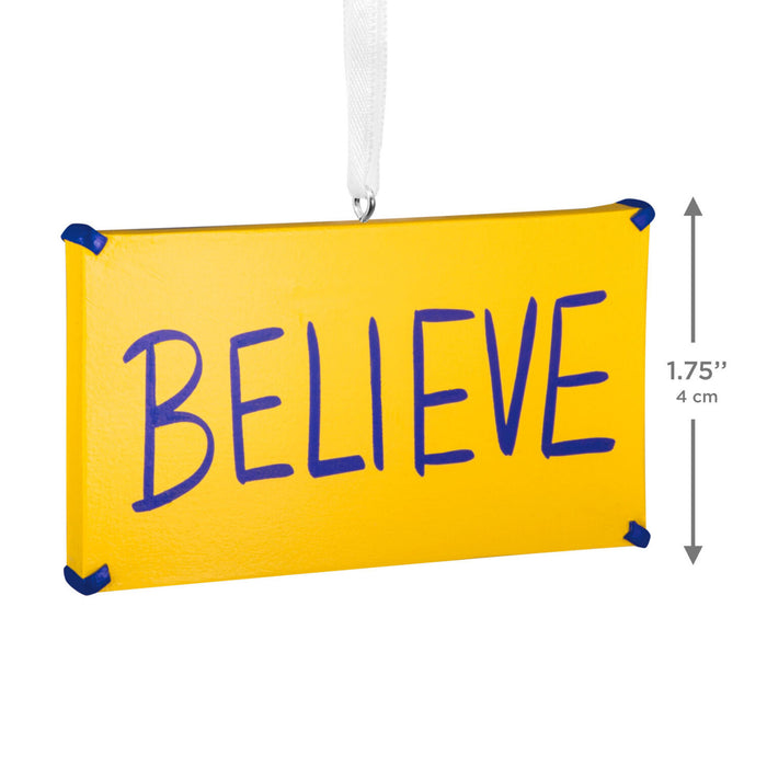 A yellow rectangular hanging sign with the word “BELIEVE” in large blue handwritten-style letters. A white hanging ribbon is attached at the top, and a vertical dimension marker on the right reads 1.