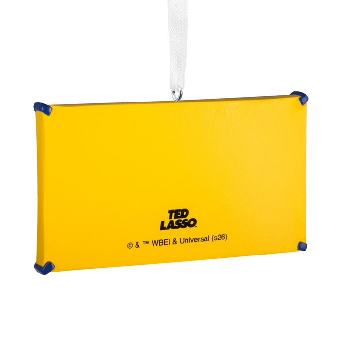 A yellow rectangular hanging ornament or plaque with blue corner protectors and a white hanging ribbon. The front is printed with the text 'TED LASSO' and smaller copyright text near the bottom, shown on a plain white background.