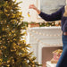 Person decorating a Christmas tree with lights and ornaments in a festive living room.