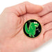 A small round enamel pin featuring a green cartoon dinosaur on a black background with a gold border, held in the center of a person's hand.