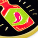 A close-up of a circular enamel pin featuring a stylized chili pepper bottle design with red, lime green, and pink colors outlined in gold on a black background.