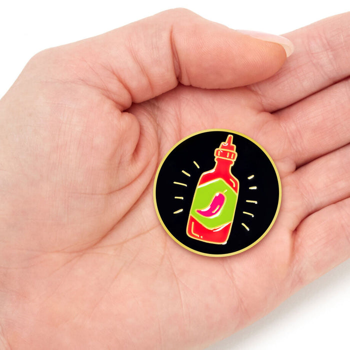 A circular enamel pin shaped like a hot sauce bottle is displayed on a person's palm. The pin features a black background with a yellow outline, a red and green bottle graphic, and a pink chili pepper illustration.