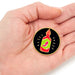 A circular enamel pin shaped like a hot sauce bottle is displayed on a person's palm. The pin features a black background with a yellow outline, a red and green bottle graphic, and a pink chili pepper illustration.