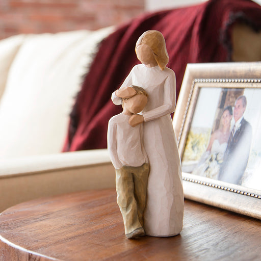 Decorative figurine of a woman holding a child on a wooden table with a blurred photo frame in the background.