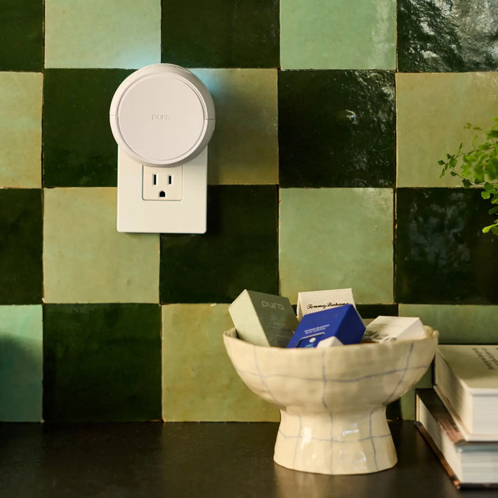 A white circular smart plug device with a round top, branded 'Karma', mounted on a green checkered tile wall above a dark countertop with a ceramic bowl and books.