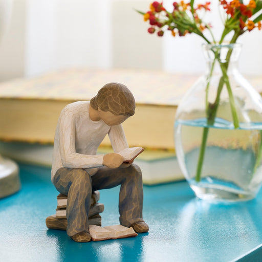 Wooden figurine of a person reading a book on a table with a vase of flowers in the background.
