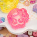A translucent pink paw-shaped gel item with embedded glitter, displayed on a white tray with pastel props and a keyboard in the background.