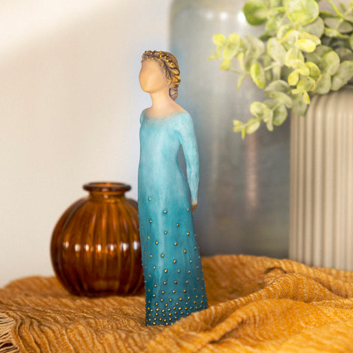 Standing figure with arms clasped behind back, hair and deep blue ombré dress are dotted with gold leaf. Figure has lighter skin and hair color

