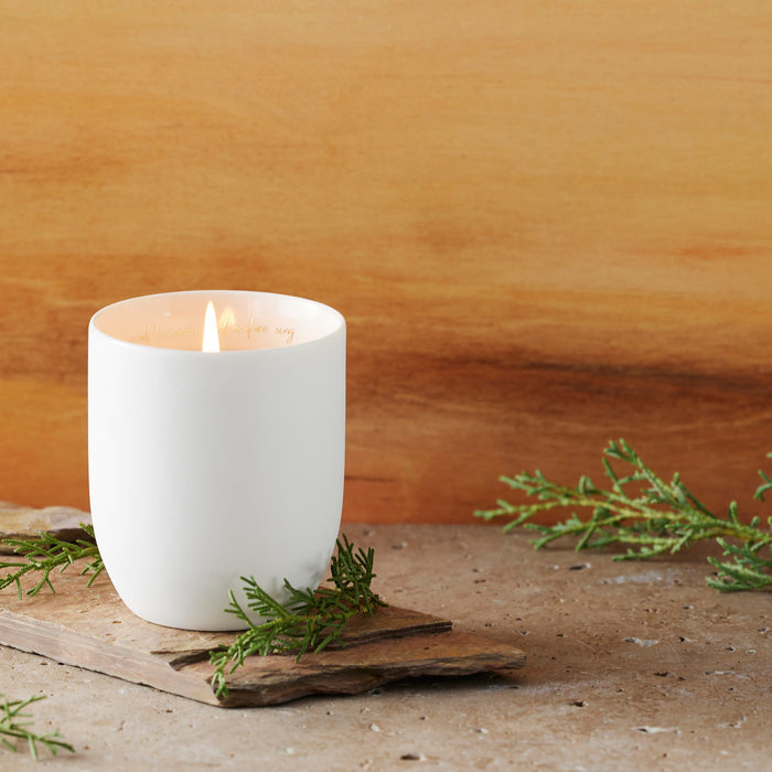 White candle in a ceramic holder on a wooden surface with greenery, against a wooden background