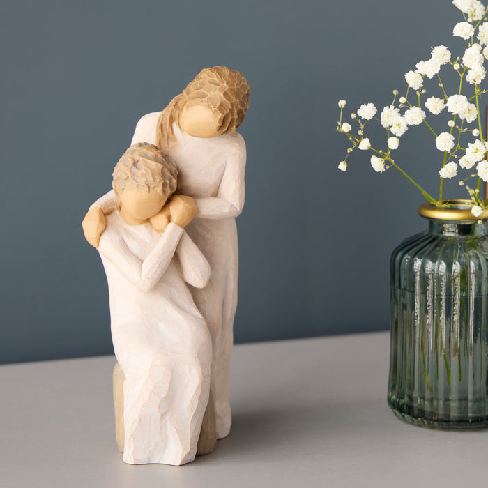 Statue of a mother and child embracing next to a vase with white flowers on a gray surface.