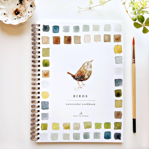 A spiral-bound watercolor workbook titled 'BIRDS' by Emily Lee Studio, featuring a watercolor bird illustration in the center and a border of watercolor color swatches. The workbook is displayed on a white surface with a paintbrush, a small dish, and green leaves.